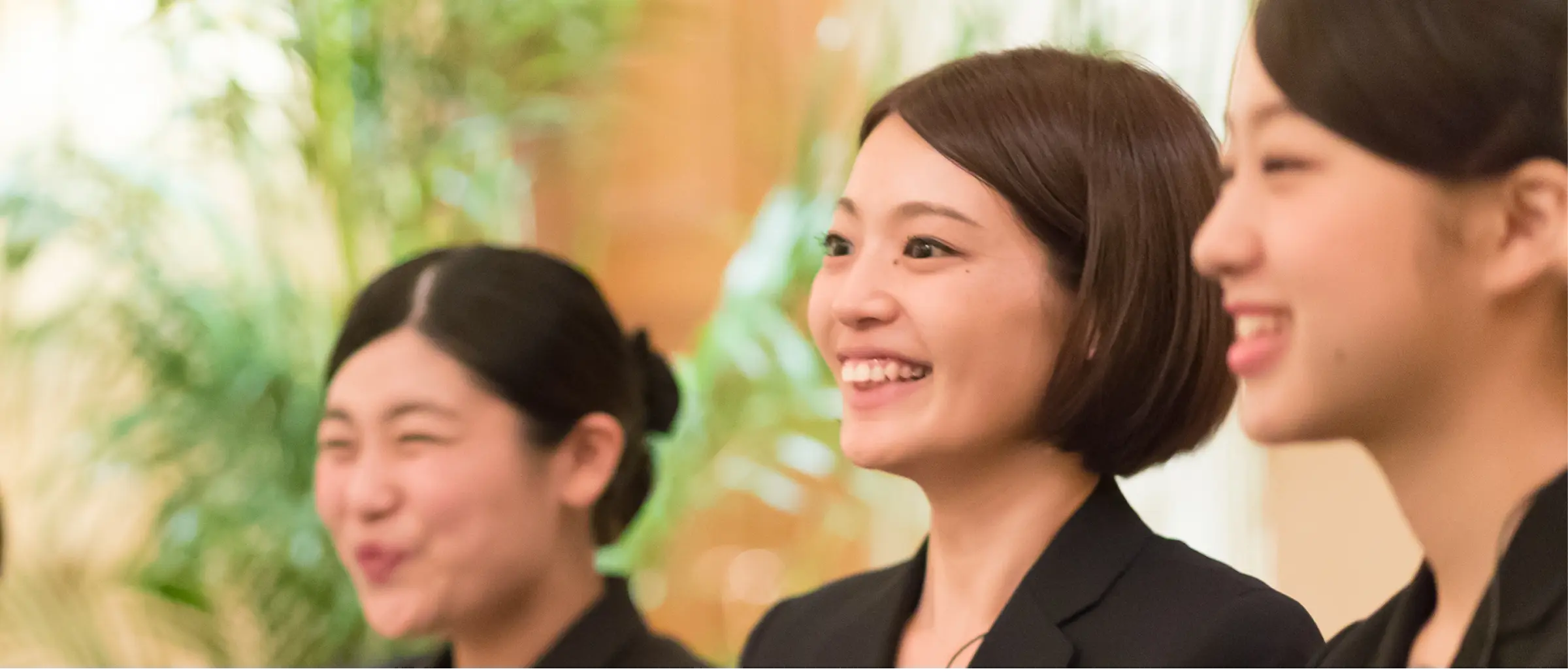 黒いスーツ姿の若い女性三人が横並びで、右方向を見ながら笑顔を見せている様子。中央のボブヘアの女性がくっきり写り、背景は観葉植物と暖かい照明がぼけた室内。