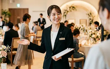 披露宴会場で、紺のスーツにイヤホンとクリップマイクをつけた女性スタッフがクリップボードを抱え、右側の相手に向けて説明するように身振りしている様子。奥には白いウェディングドレスの花嫁がぼけて写る。