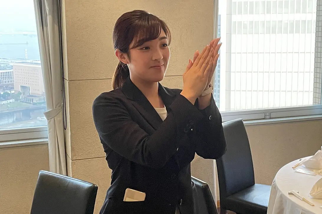 会議室で、スクリーンの前に座り、穏やかに微笑みながら話を聞いている女性