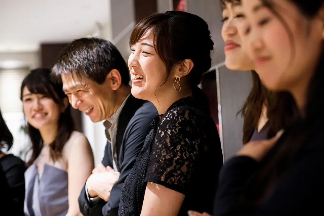 親睦会のような会場で、横並びになり笑い合う複数人。中央の女性が笑顔で会話に参加している