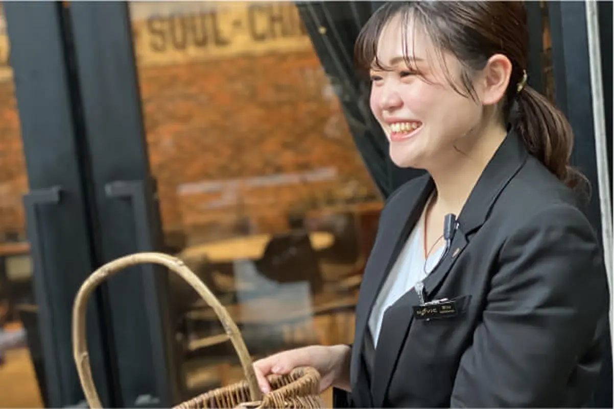 名札に「nOvic」「Producer」とある女性が、カゴを持ち、ガラス扉のあるビル入口の前に立っている