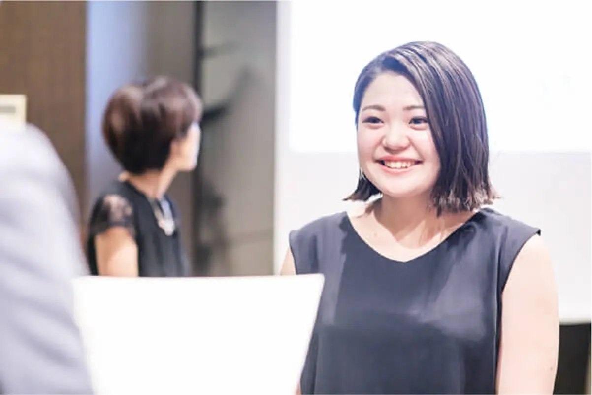 長い黒髪の女性が、明るい室内で笑顔になりながら会話している。黒のトップスとチョーカーを着用
