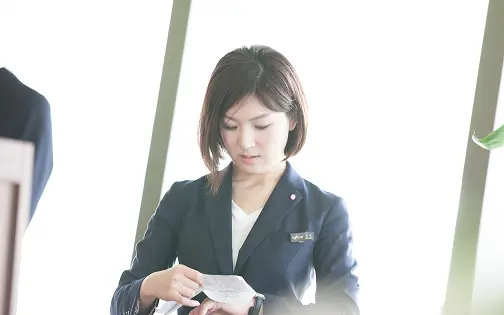 街中で黒いタクシーからストラップレスの白いウェディングドレスの花嫁が降りる瞬間。ピンクのバラのブーケを持ち、紺のスーツの男性が手伝い、黒いビジネススーツの女性が笑顔で迎えている。