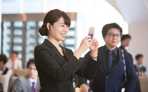 黒いスーツを着た二人の女性スタッフが、ヘッドセットをつけたまま書類やパンフレットを一緒に確認している様子。壁は石目調、横に花のアレンジメント。