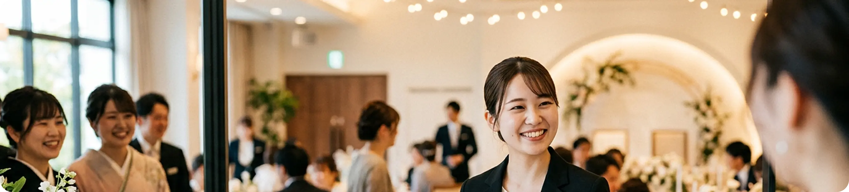 電飾の明るい披露宴会場で、スーツ姿の女性スタッフが笑顔でゲストを案内している様子。奥には円卓と花のアーチが見える
