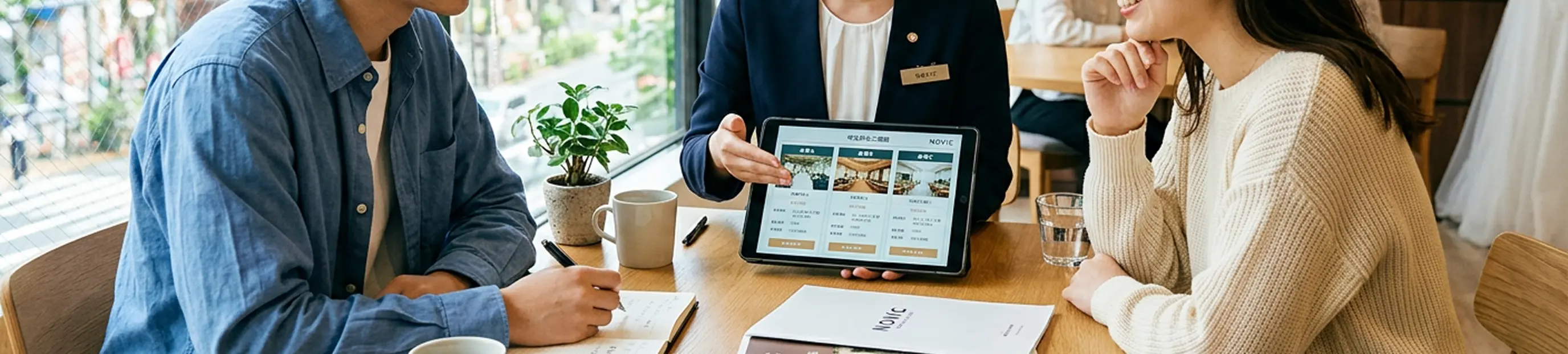 「NOVIC WEDDING SALON」のロゴが掲げられた明るい店内で、紺のスーツの女性スタッフがタブレットで会場案内をし、若いカップルがノートにメモを取りながら相談を受けている様子。テーブルにはコーヒー、水、NOVICのパンフレットが並ぶ。窓の外には街路樹と建物が見える。