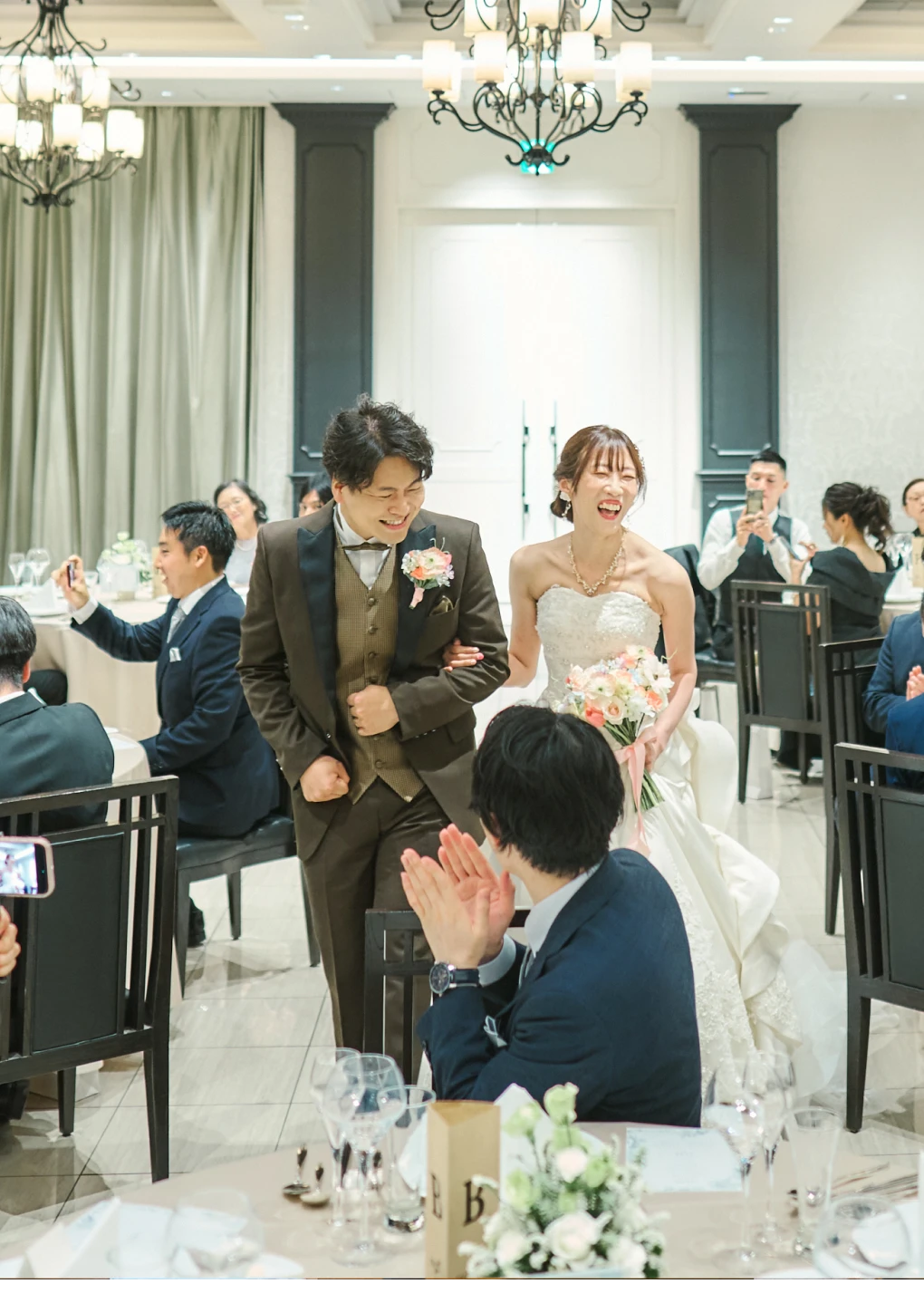 明るい披露宴会場で、茶色のスリーピースにピンクのブートニアの新郎と、ストラップレスの白いレースドレスにピンク系ブーケの花嫁が並んで歩きながら笑い、周囲のゲストが拍手やスマートフォン撮影で迎える様子。天井にはシャンデリア、奥には白い二重扉。