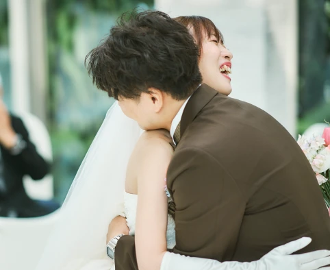 ストラップレスの白いウェディングドレスにベールと肘丈グローブの花嫁が、茶色のスーツの新郎に抱きつき、目を閉じて大声で笑っているクローズアップ。背景は明るくぼけ、緑の木々が見える。