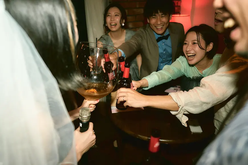 レンガ壁の室内パーティーで、ベールをつけた花嫁が特大のワイングラスとマイクを持ち、若い男女数名が笑顔でビール瓶をグラスに合わせて乾杯しようとしている様子。テーブルにはスマートフォンや紙類。