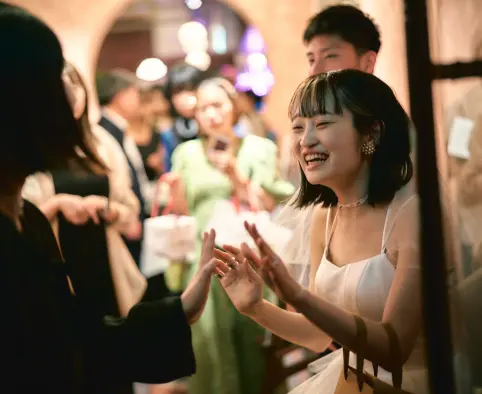 披露宴のような会場で、白いキャミソール型のドレスにパールのアクセサリーの女性が目を細めて笑い、両手を前に伸ばして誰かと触れ合おうとしている様子。背景には正装の列席者がぼけ、奥に紫と白の照明のアーチが見える。