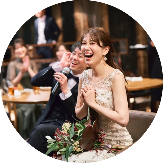 円形に切り抜かれた写真。ビーズと花柄の白いノースリーブドレスの花嫁と、タキシードでマイクを持つ新郎が並んで座り、二人とも笑い、花嫁は手を胸前で組み、膝上に深紅のブーケ。背景では列席者が拍手している。