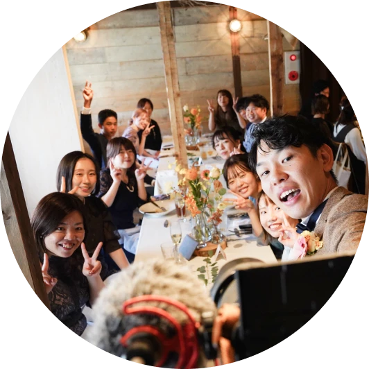 円形に切り抜かれた俯瞰気味の写真。木目の壁の会場で長いテーブルに座る若い男女約十人がカメラに向かって笑い、複数人がピースサイン。白いテーブルクロスに桃色の小花、シャンパングラス。手前には映像用カメラとマイクのウィンドシールドが写る。