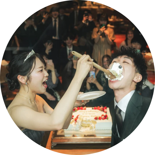 円形に切り抜かれた写真。ティアラとダークカラーのストラップレスドレスの花嫁が、大きな木のスプーンで白い生デコレーションケーキを新郎に口いっぱいに食べさせようとし、新郎は口を大きく開けて受け止めている様子。ケーキはイチゴ飾りの二段。背景のゲストがスマートフォンで撮影している。
