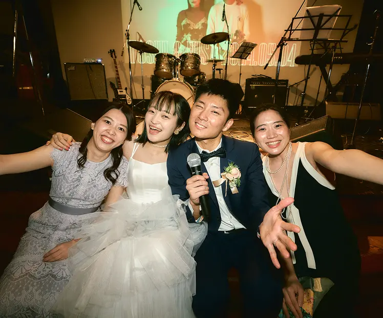 ライブステージの前で、ラベンダー色のレースドレスの女性、白いティアードのウェディングドレスの女性、紺のスーツにマイクを持つ男性、黒いドレスにパールネックレスの女性の四人が寄り添い、カメラに向かって笑顔で写る様子。背景にはドラムやキーボードなどの楽器と、投影された「TOSHI & … 2024.05.04」と書かれた映像。