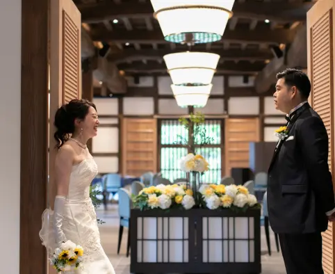 木製のルーバー扉の間から見た披露宴会場で、肘丈グローブと白いウェディングドレスの花嫁が白とひまわりのブーケを持ち微笑み、向かいには黄色のひまわりのブートニアをつけたタキシードの新郎が立っている様子。天井には白いボウル型のペンダントライト、奥には花飾りと青い椅子の席が見える。
