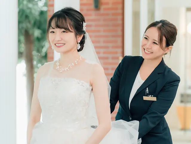 レンガ壁の前で、パールの髪飾りとチュールベールのノースリーブの白いウェディングドレスの花嫁が微笑み、後ろから紺のスーツの女性スタッフがドレスの背中やベールを整えている様子。