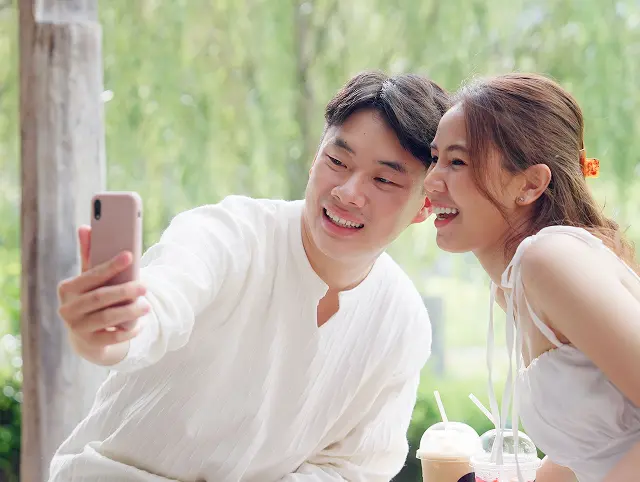 屋外のテーブルで、白いリブシャツの男性がスマートフォンで二人の自撮りをし、白いキャミソールの女性が寄り添って笑っている様子。手前のテーブルにはアイスドリンクが二杯、背景は緑の木々がぼけた明るい昼下がり。