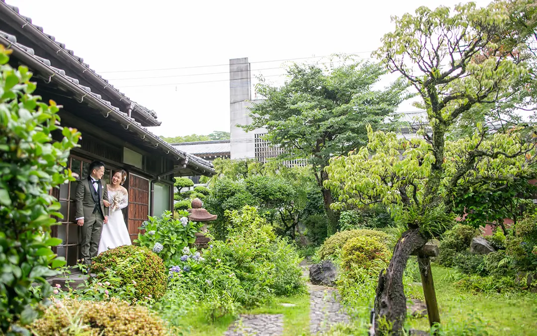 緑豊かな日本庭園の小道に、白いウェディングドレスの花嫁とダークスーツの新郎が寄り添って立っている様子。背後には古民家風の建物の軒先、石燈篭や青いアジサイが咲く庭が広がる。