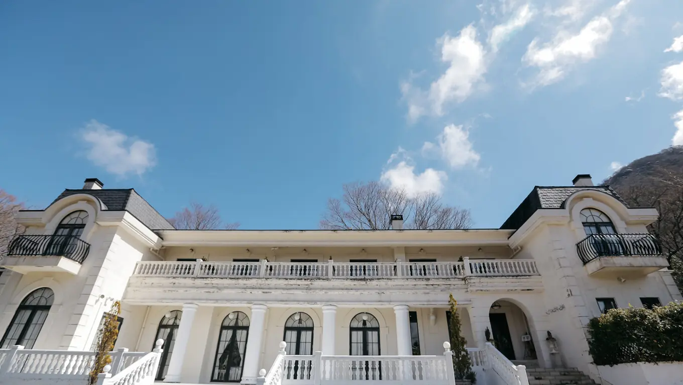 青空を背景に、低い視点から見上げた白い外壁の二階建て洋館。アーチ型の大きな窓と扉、石造りの手すりのテラス、二階のバルコニー、両脇の出窓に小さな鉄製バルコニー、濃い色の瓦屋根。奥には落葉樹と山の斜面が見える。