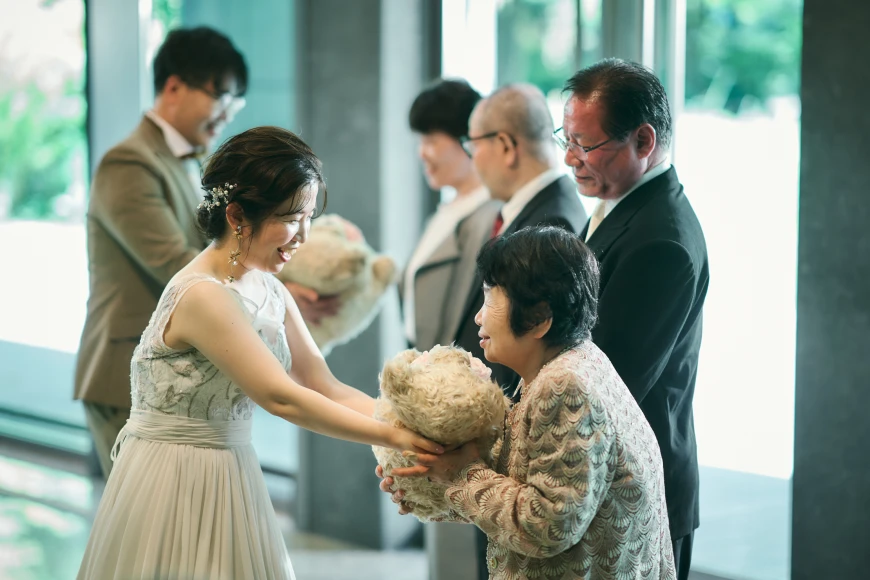 明るい式場で、パールの髪飾りとビーズ装飾の白いウェディングドレスの花嫁が、大きな薄茶色のぬいぐるみのクマを、薄色の柄ジャケットの高齢女性に手渡している様子。奥ではベージュのスーツの新郎も別の列席者に同様のクマを渡している。