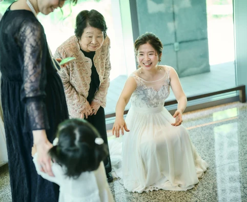 大理石調の床の明るいロビーで、銀灰色の花柄刺繍のノースリーブの白いウェディングドレスの花嫁がひざまずき、両手を広げて幼い子どもに笑顔で手を伸ばしている様子。子どものそばにはベージュのレースのジャケットの高齢女性と、黒いレースのドレスの女性。背景は大きな窓と緑。
