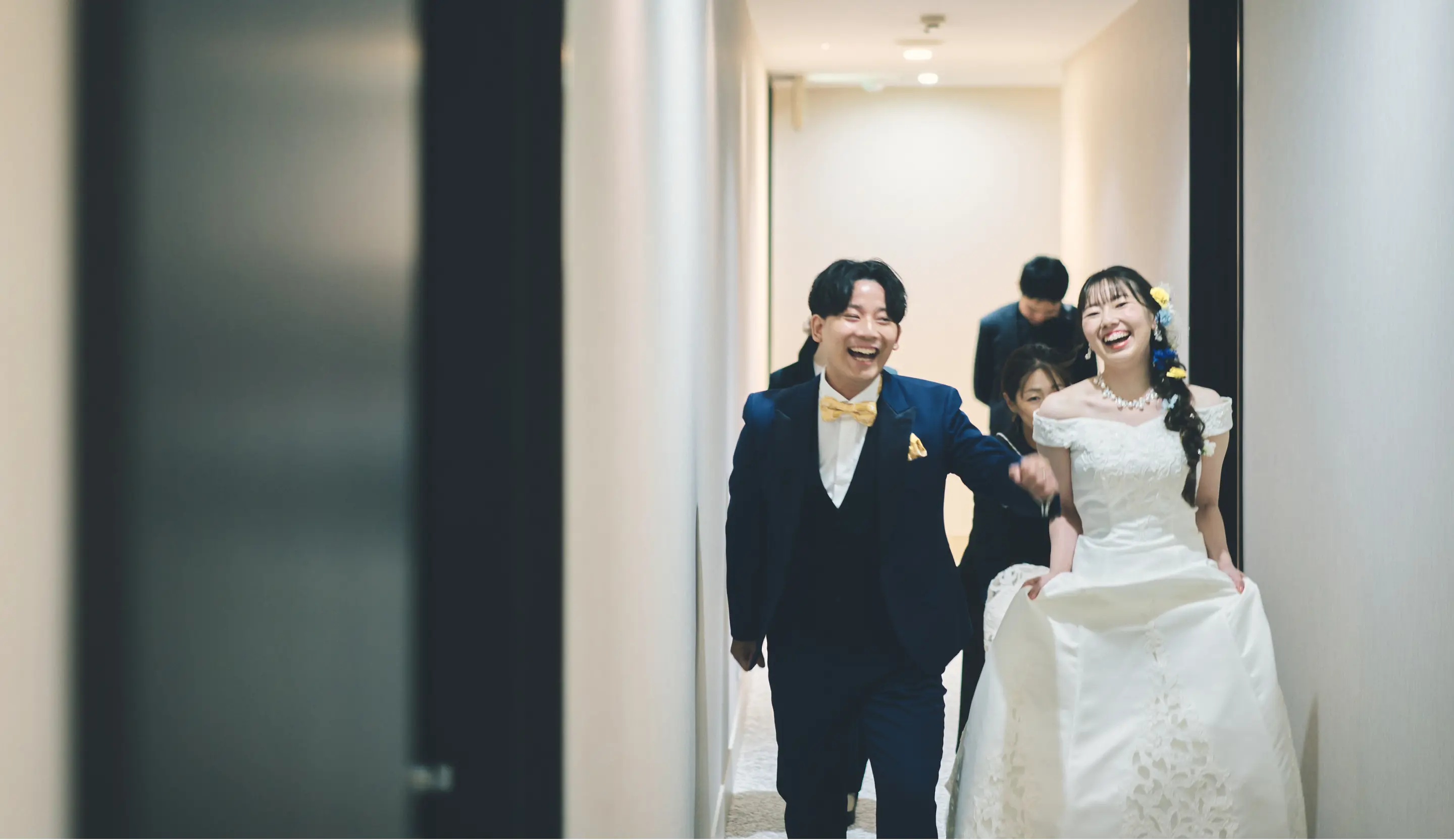 結婚式の廊下を歩く新郎新婦。ネイビーのタキシードに黄色の蝶ネクタイの新郎と、オフショルダーの白いウェディングドレスに髪飾りをつけた新婦が、笑顔でカメラ方向へ歩いている様子。