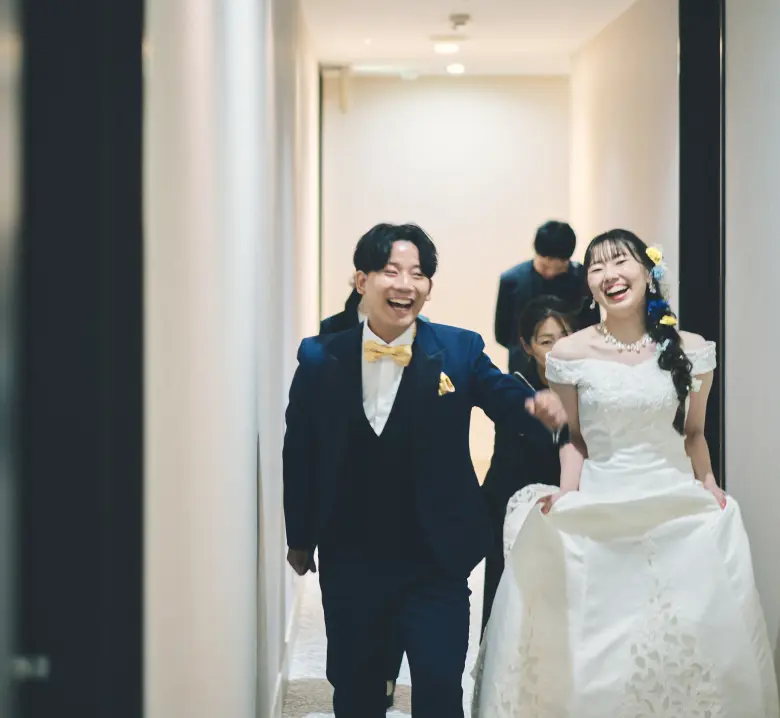 結婚式の廊下を歩く新郎新婦。ネイビーのタキシードに黄色の蝶ネクタイの新郎と、オフショルダーの白いウェディングドレスに髪飾りをつけた新婦が、笑顔でカメラ方向へ歩いている様子。
