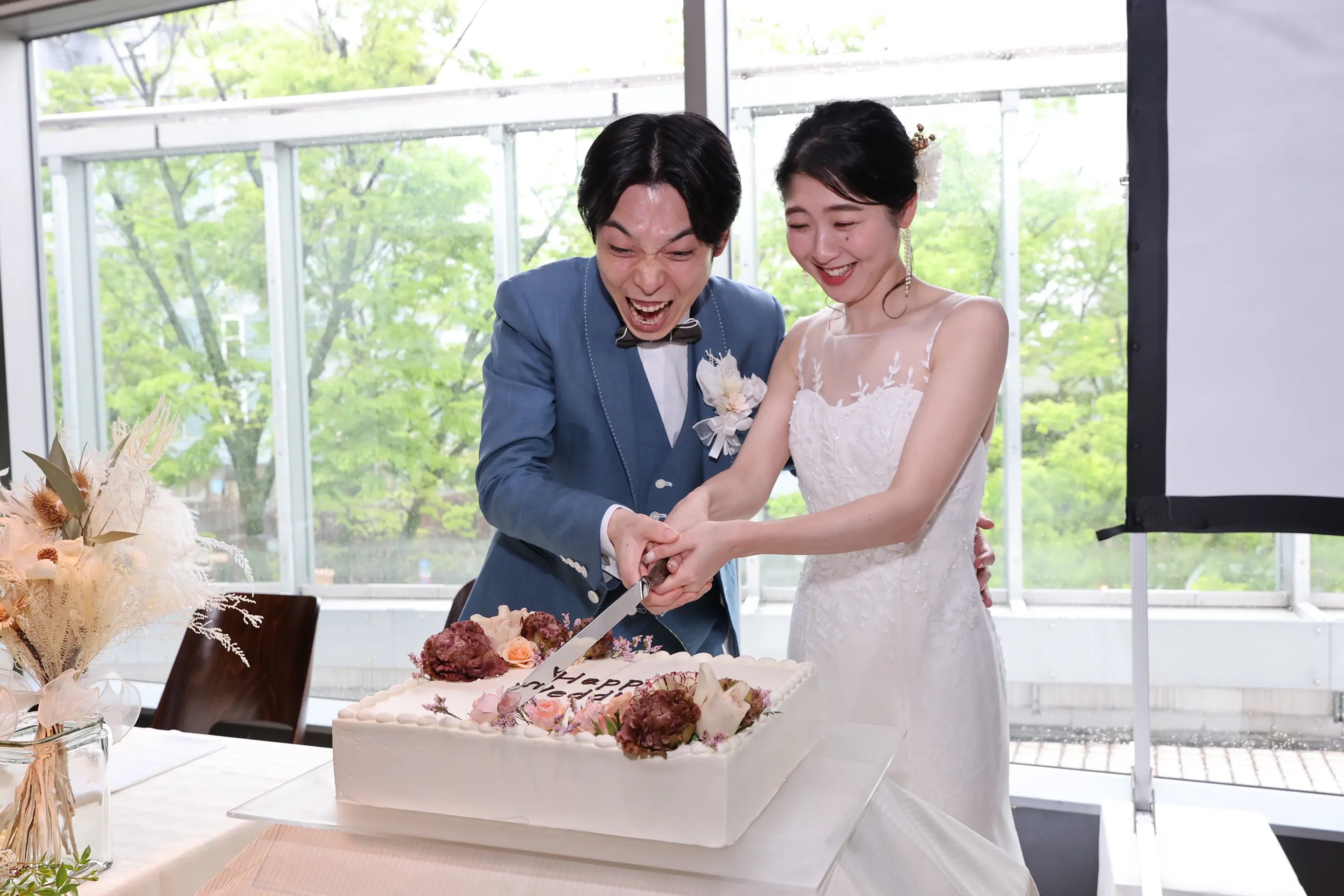 披露宴で、青みがかったグレーのスーツの新郎と白いレースのウェディングドレスの花嫁が、二人でナイフを入れて角型のウェディングケーキをカットしている様子。窓の外には緑の木々が見える。