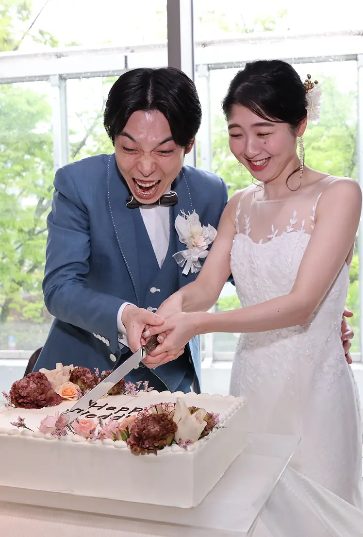 披露宴で、青みがかったグレーのスーツの新郎と白いレースのウェディングドレスの花嫁が、二人でナイフを入れて角型のウェディングケーキをカットしている様子。窓の外には緑の木々が見える。