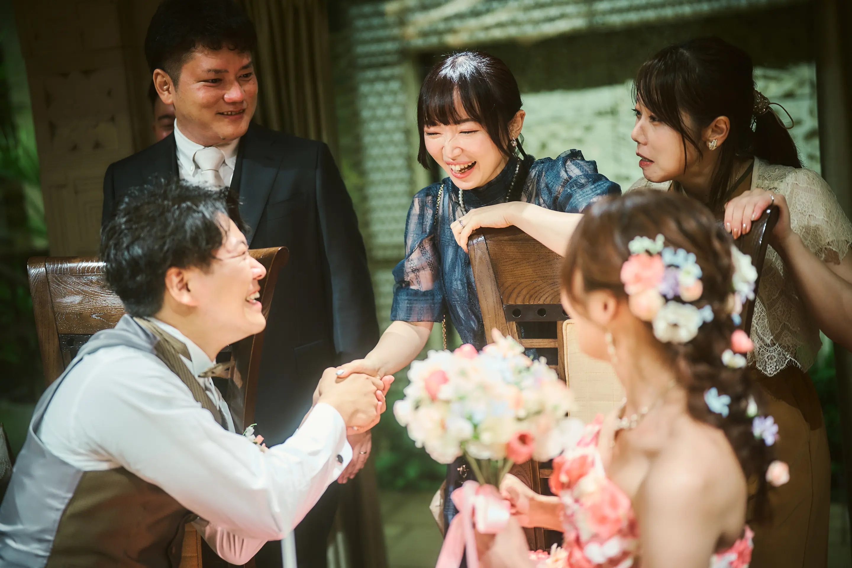 披露宴のテーブル席で、茶色のベストと蝶ネクタイの新郎が女性ゲストと握手し笑い合い、花嫁は小花飾りのついた髪型とピンクのストラップレスドレスで背中越しに写っている様子。周囲のゲストも笑顔。