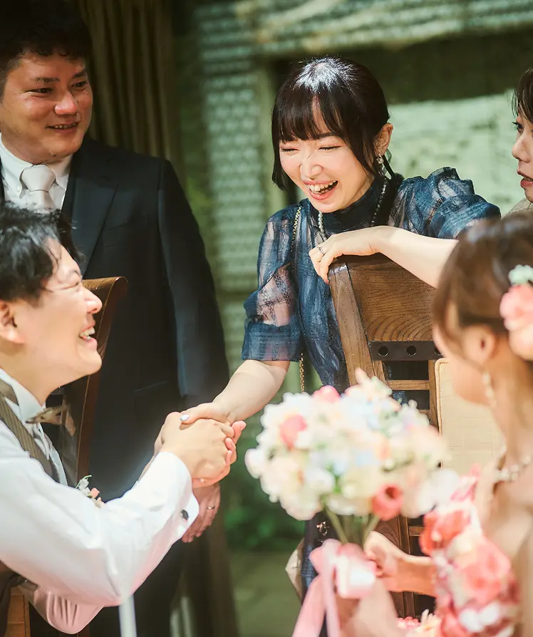 披露宴のテーブル席で、茶色のベストと蝶ネクタイの新郎が女性ゲストと握手し笑い合い、花嫁は小花飾りのついた髪型とピンクのストラップレスドレスで背中越しに写っている様子。周囲のゲストも笑顔。