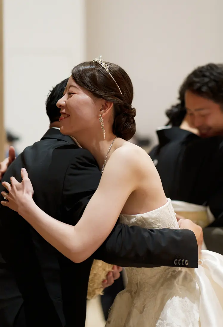 披露宴の会場で、ティアラとストラップレスの白いレースドレスの花嫁が黒いスーツの男性と抱擁し、奥ではタキシードの新郎が金色の帯の黒留袖の女性と抱擁している様子。