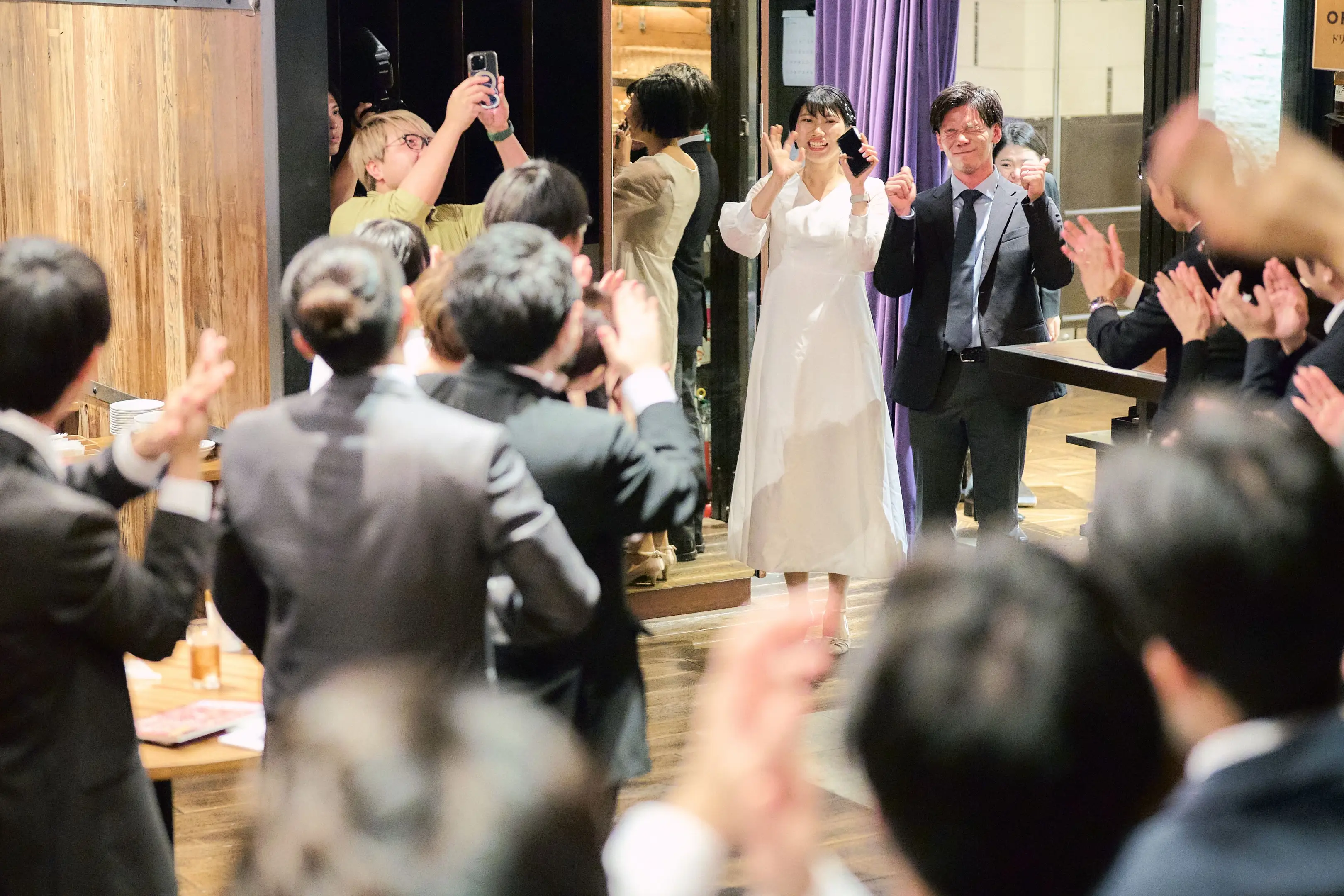 披露宴のような会場で、白いキャミソール型のドレスにパールのアクセサリーの女性が目を細めて笑い、両手を前に伸ばして誰かと触れ合おうとしている様子。背景には正装の列席者がぼけ、奥に紫と白の照明のアーチが見える。