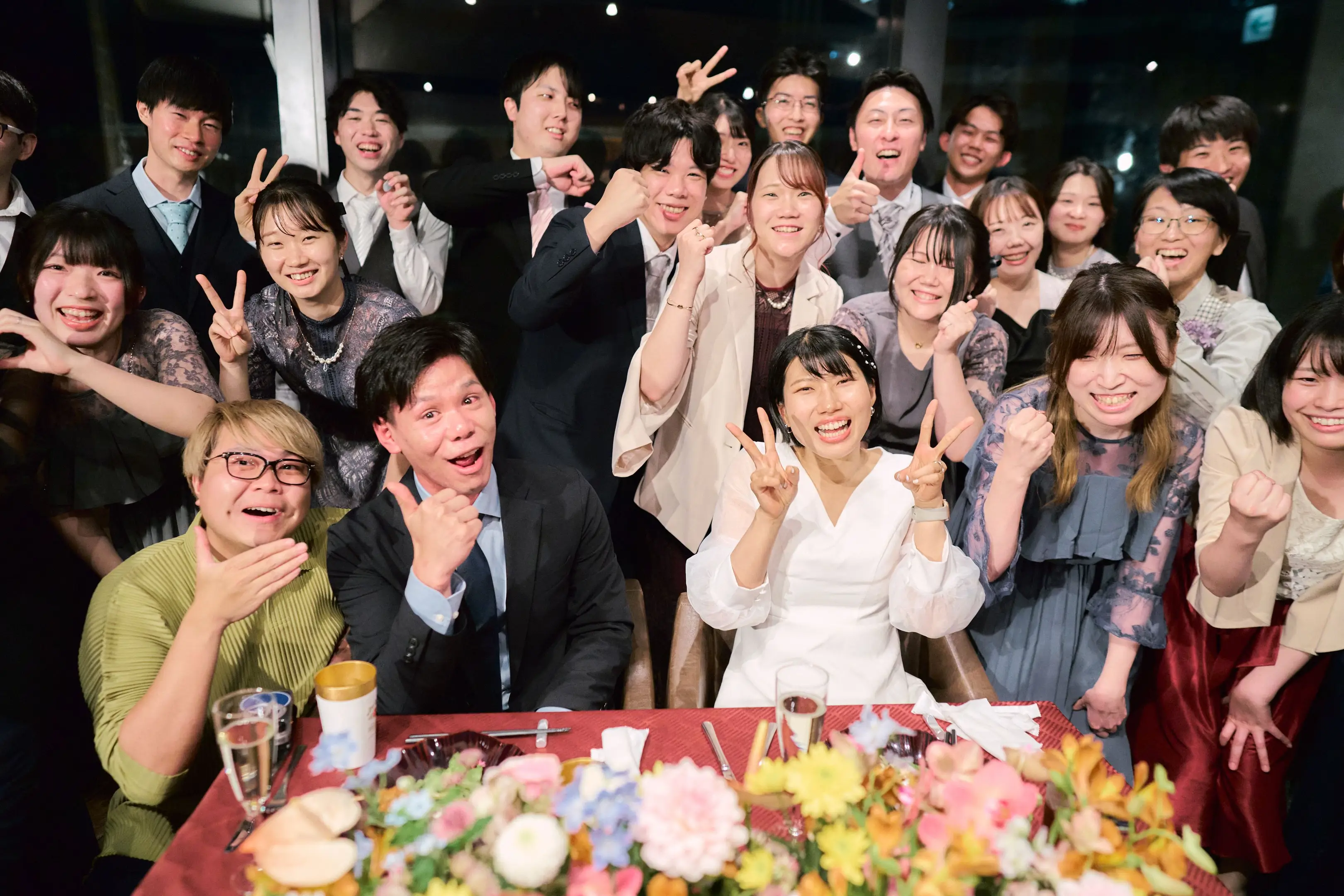 レンガ壁の室内パーティーで、ベールをつけた花嫁が特大のワイングラスとマイクを持ち、若い男女数名が笑顔でビール瓶をグラスに合わせて乾杯しようとしている様子。テーブルにはスマートフォンや紙類。