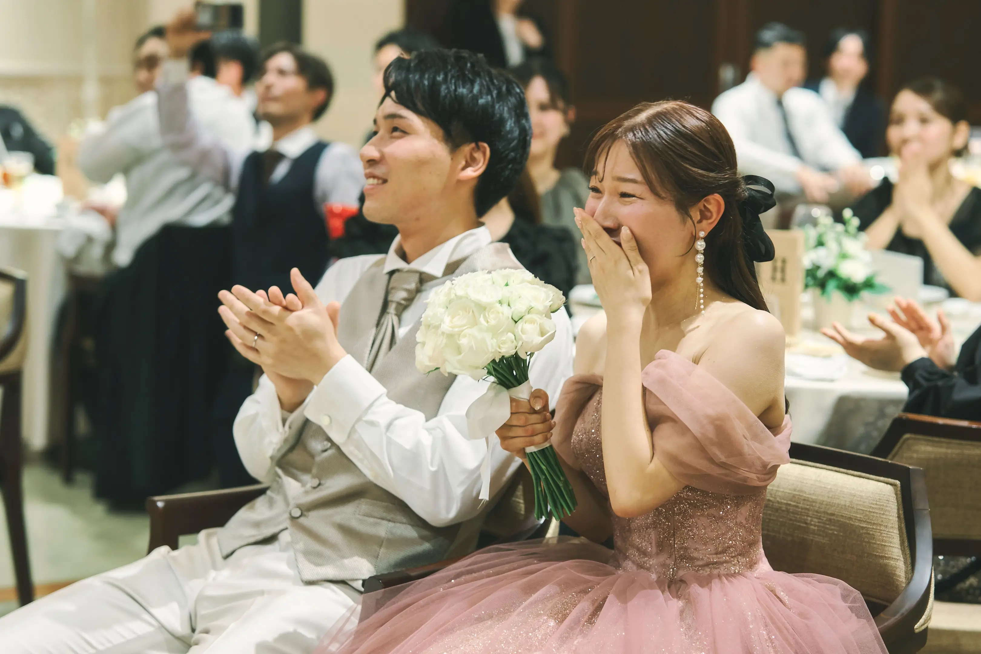 円形に切り抜かれた写真。ビーズと花柄の白いノースリーブドレスの花嫁と、タキシードでマイクを持つ新郎が並んで座り、二人とも笑い、花嫁は手を胸前で組み、膝上に深紅のブーケ。背景では列席者が拍手している。