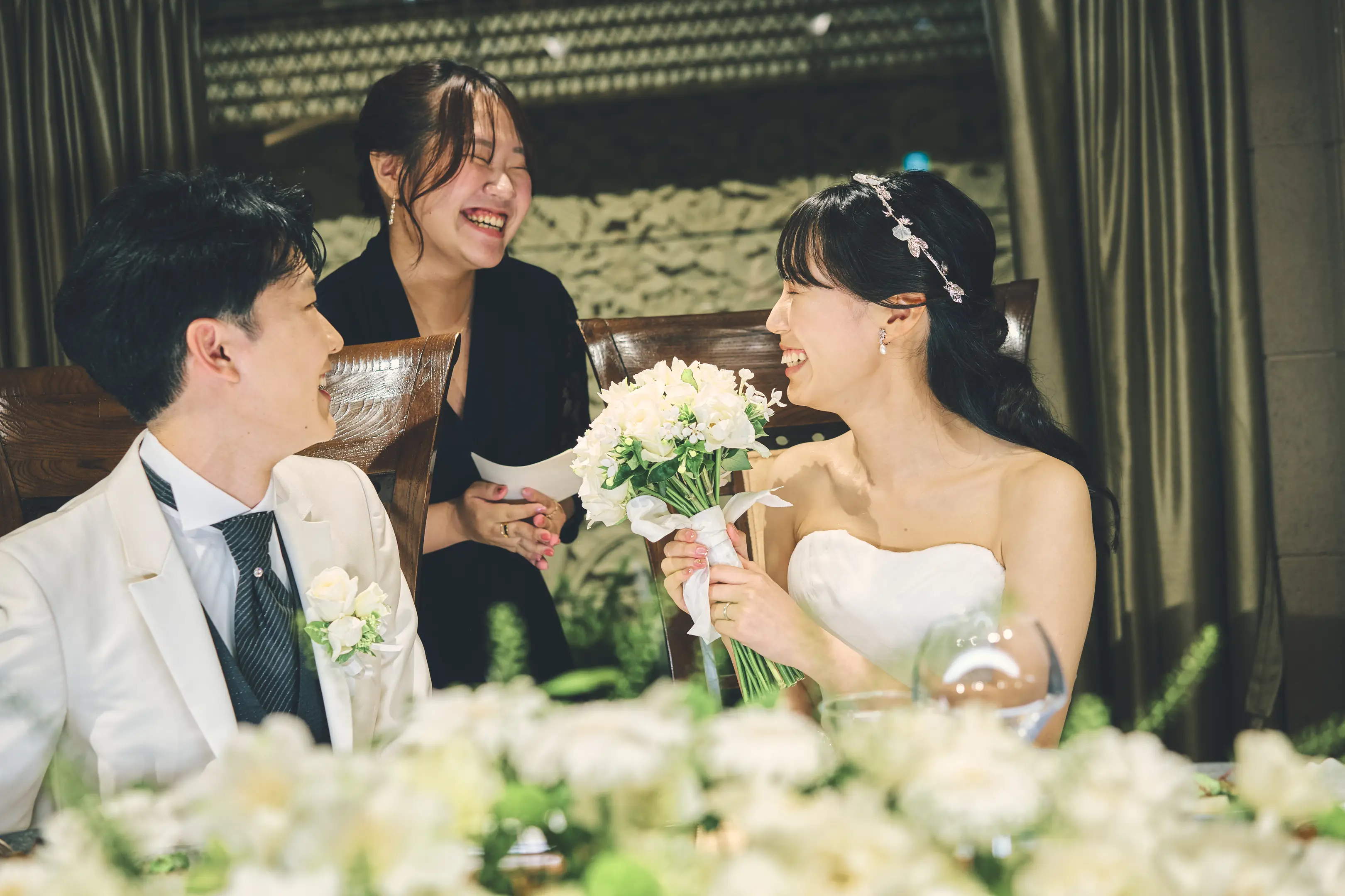 披露宴のテーブル席で、茶色のベストと蝶ネクタイの新郎が女性ゲストと握手し笑い合い、花嫁は小花飾りのついた髪型とピンクのストラップレスドレスで背中越しに写っている様子。周囲のゲストも笑顔。