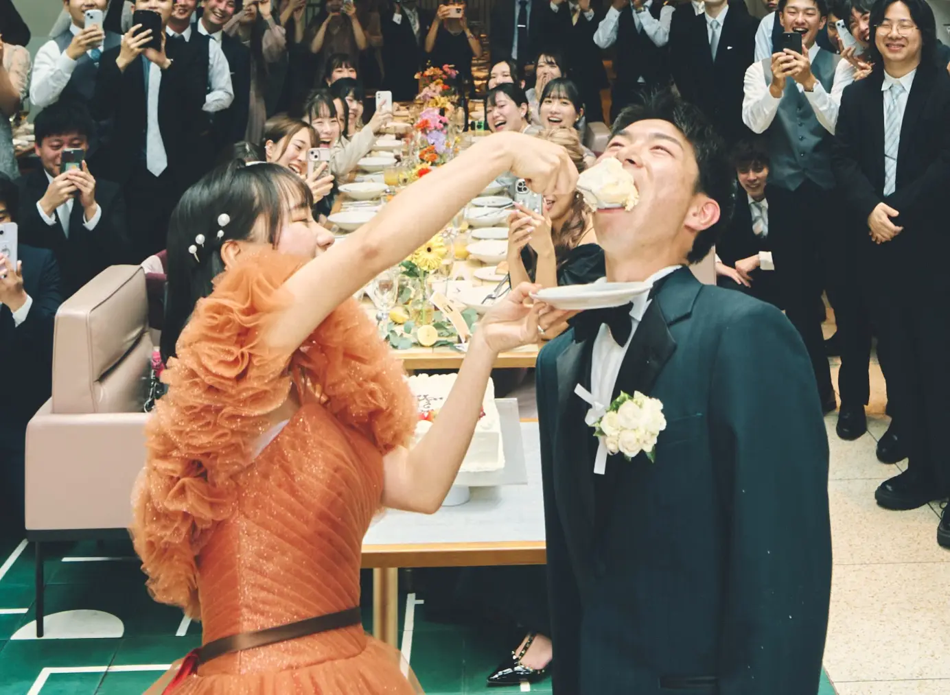 円形に切り抜かれた写真。ティアラとダークカラーのストラップレスドレスの花嫁が、大きな木のスプーンで白い生デコレーションケーキを新郎に口いっぱいに食べさせようとし、新郎は口を大きく開けて受け止めている様子。ケーキはイチゴ飾りの二段。背景のゲストがスマートフォンで撮影している。